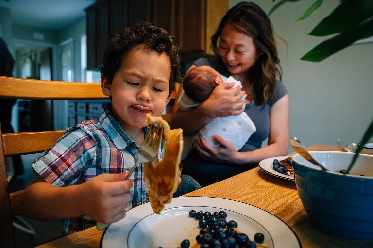 Mother holds and smiles at newborn baby while sitting at kitchen table eating breakfast. Little boy picks up pancake with his fork.