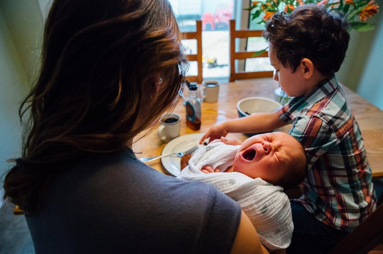 Mother sits and eats breakfast at kitchen table with her son while holding newborn baby boy.