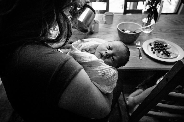 Black and white. Mother eats breakfast while holding newborn baby boy.