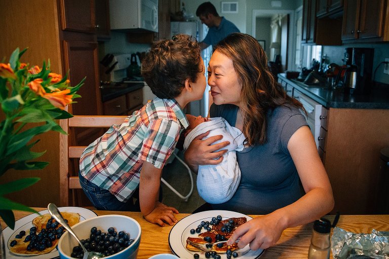 Mother holding newborn baby kisses son as they sit eating breakfast.