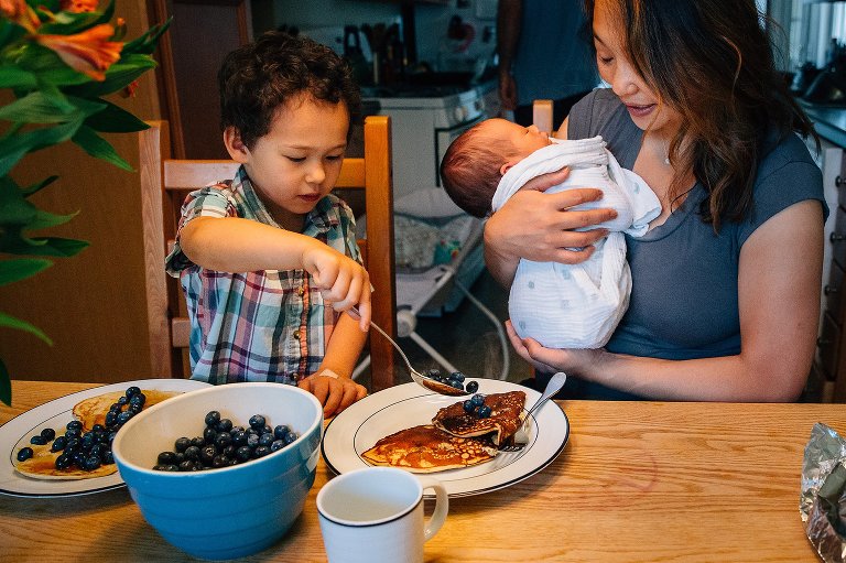 Mother holding newborn baby at kitchen table, son adds blueberries onto plate of pancakes.