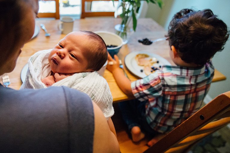 Mother holding newborn baby at kitchen table with her son sitting next to her eating pancakes and blueberry.