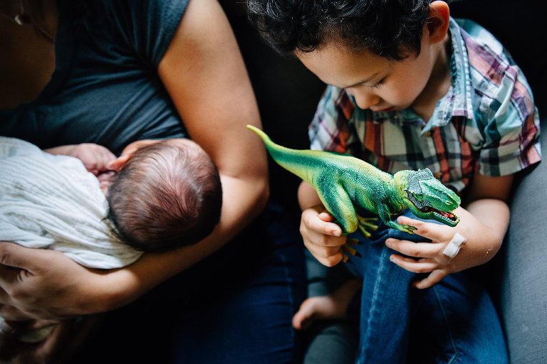 Mother sitting on couch breastfeeding newborn baby. Little boy sitting on the couch next to her playing with dinosaur toy.