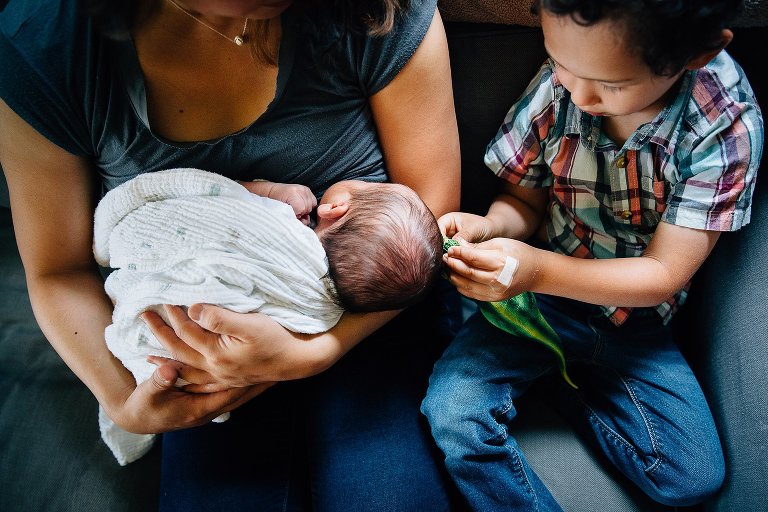 Mother sitting on couch breastfeeding newborn baby. Little boy sitting on the couch next to her playing with dinosaur toy.