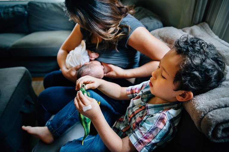 Mother sitting on couch breastfeeding newborn baby. Little boy sitting on the couch next to her playing with dinosaur toy.