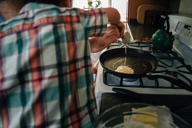 Father helps son pour pancake batter onto hot griddle on the stovetop.