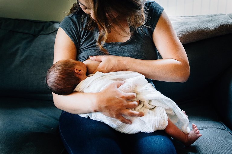 Mother sitting on couch breastfeeding swaddled newborn baby.