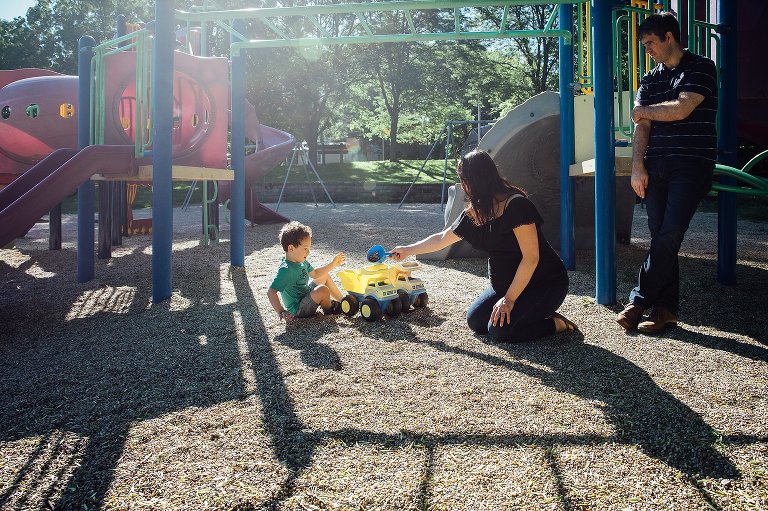 Family playing at a park. Pregnant mother kneeling on the ground playing with her son at a park while father leans against the play structure watching them.