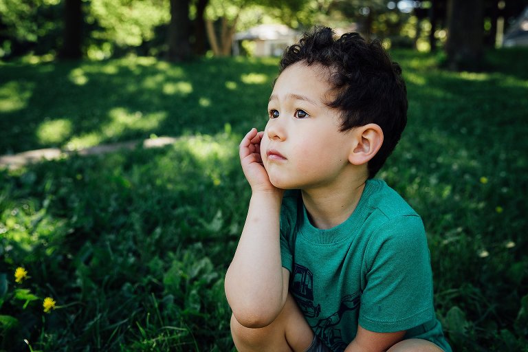 Portrait of young boy sitting in the grass resting his head on his hand looking at the trees around him.