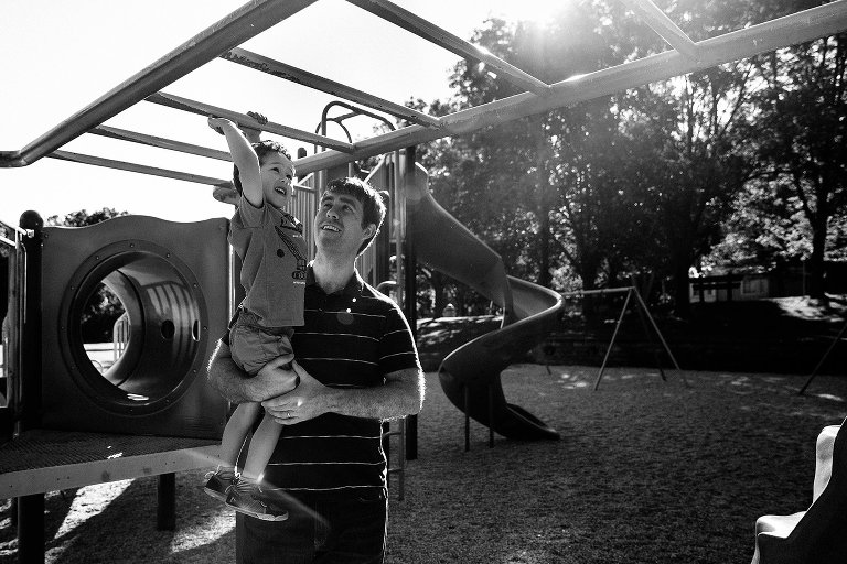 Black and white. Father helps son climb the monkey bars across park playground.