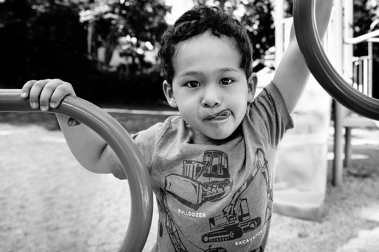 Black and white. Young boy standing and holding onto park play set licking his lips.