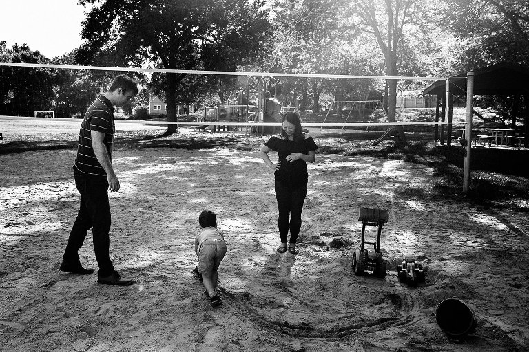 Black and white. Maternity photo of expecting mother holding her belly while watching her husband and son play in the sand at a park.