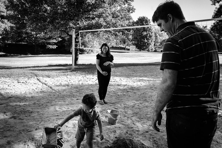 Black and white. Maternity photo of expecting mother holding her belly while watching her husband and son play in the sand at a park.