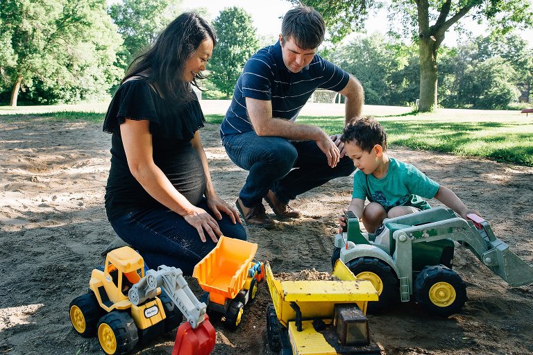 Pregnant mother, father, and son sit in the sand at a park playing with toy trucks.
