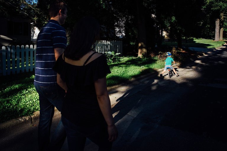 Expecting mother walks down the street holding hands with her husband watching their young son bike down the street.