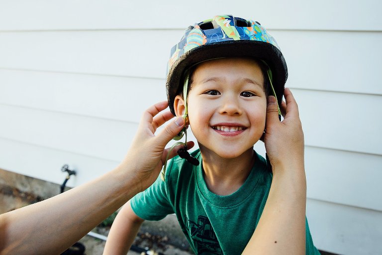 Young boy smiles as parent helps put on him helmet before going to the park as a family.