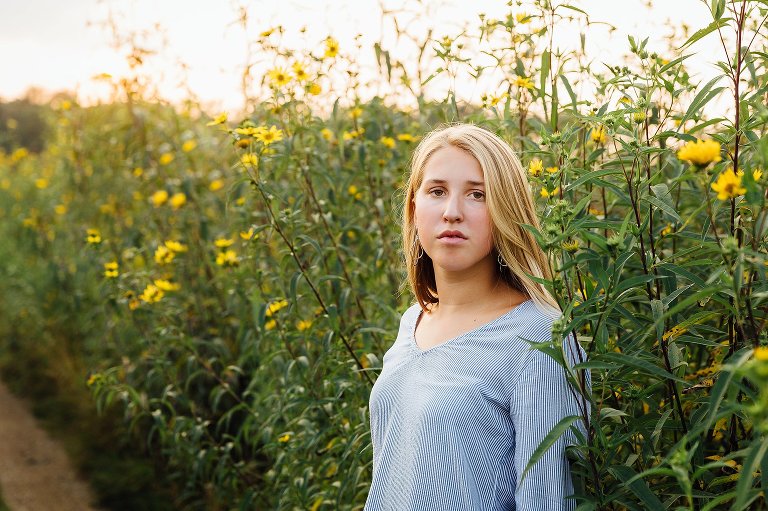 High school senior girl stands in a field of yellow flowers as the sun sets behind her. She makes eye contact with camera. 