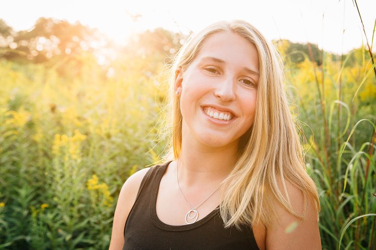 High school senior stands in field of yellow flowers with warm sun flare behind her. 