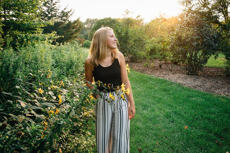 High school senior girl stands near yellow flowers as the sun sets behind her. 
