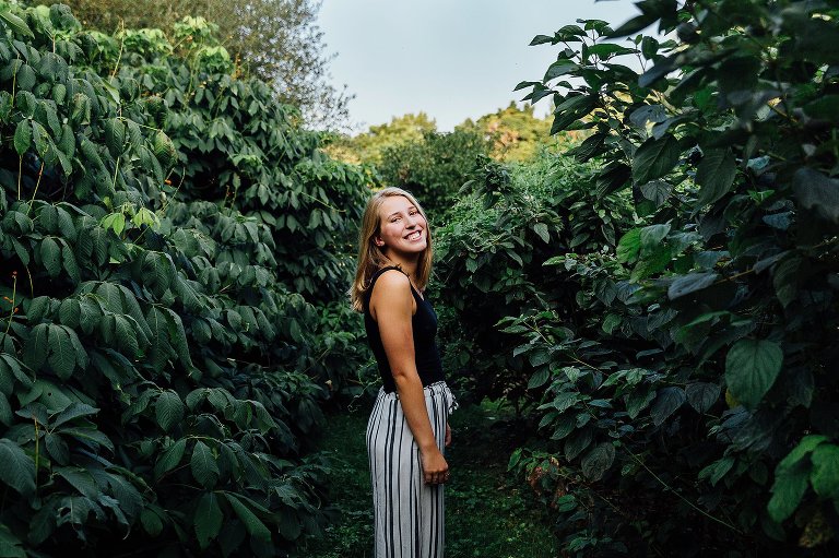 High school girl stands between two dark green bushes. Blue sky.