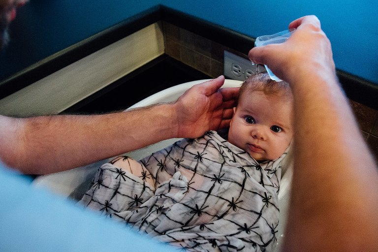 Father holds a small cup of water to infant child's head for a bath. The child is covered in a light 