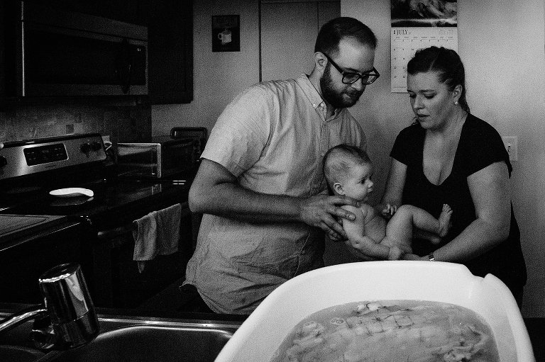Grainy black and white image. Both parents help put infant child in a bathtub on the kitchen counter. 