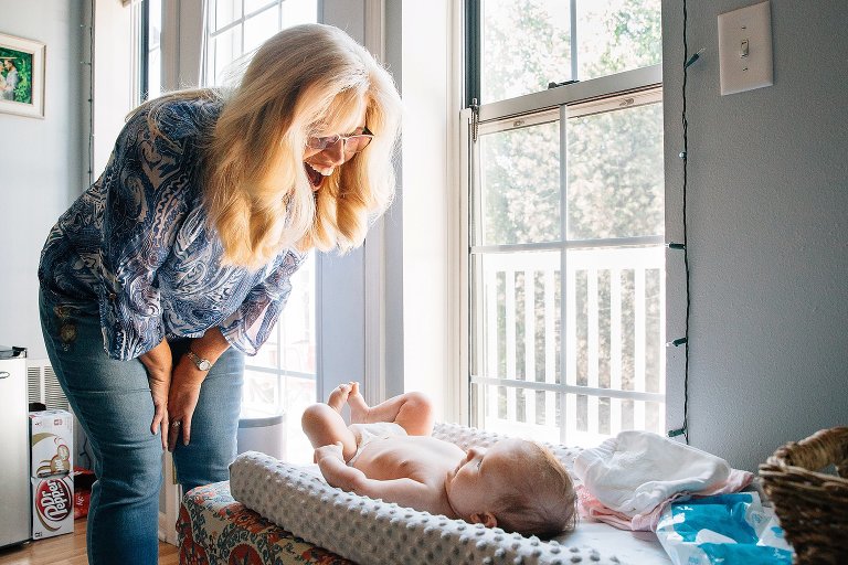 Grandma joyfully smiles with infant granddaughter as she lies on a changing pad ready for a bath. 