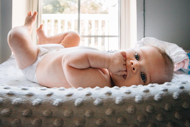 Infant child makes eyes contact with camera. She holds fist in her mouth and her feet are in the air.
