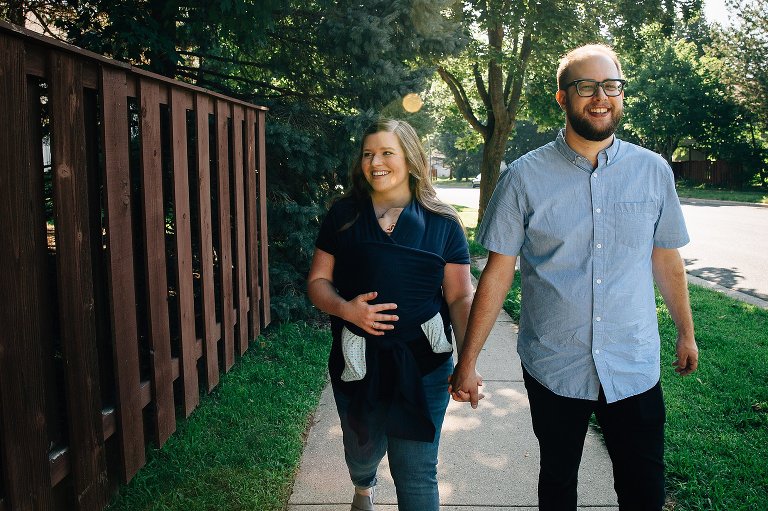 Mother and father take a walk with sleeping infant daughter being worn by the mother. The couple is holding hands and sun flare is streaming in. 