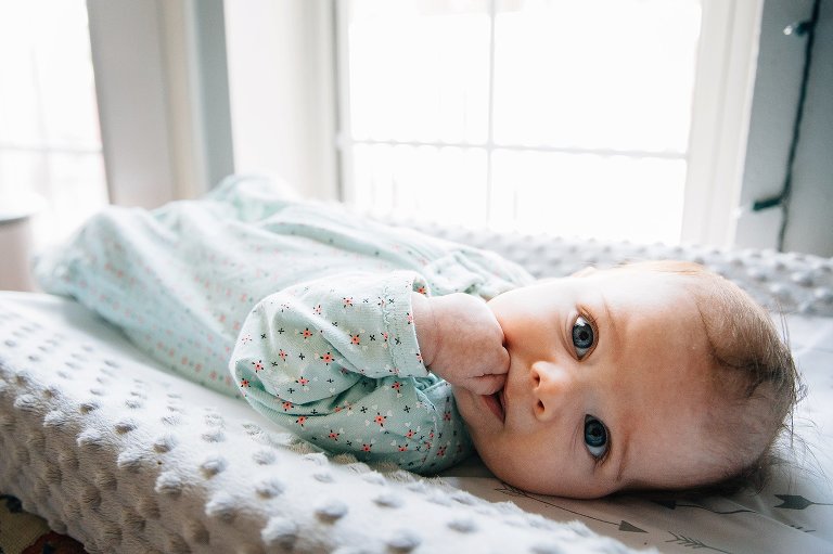 3 month old baby makes eye contact with camera, has fist in mouth, laying on a changing pad. 