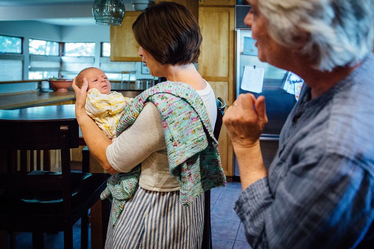 Mother holds newborn baby while standing in the kitchen. Grandma watches from behind.