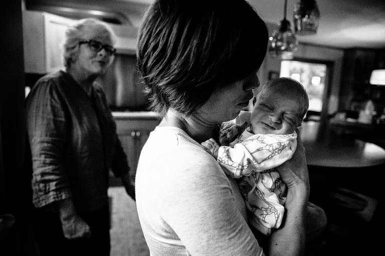 Black and white. Mom holds newborn baby in the kitchen while grandma watches.