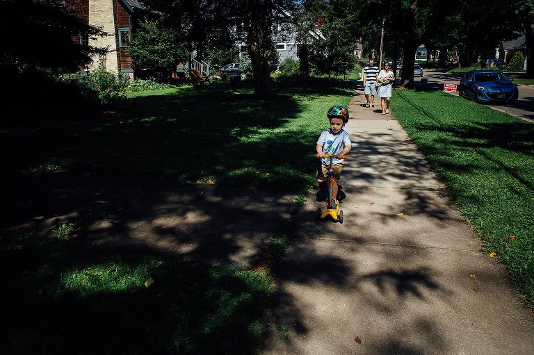Young boy scooters down the sidewalk with parents walking behind him. Shadows of the trees shine onto the sidewalks.