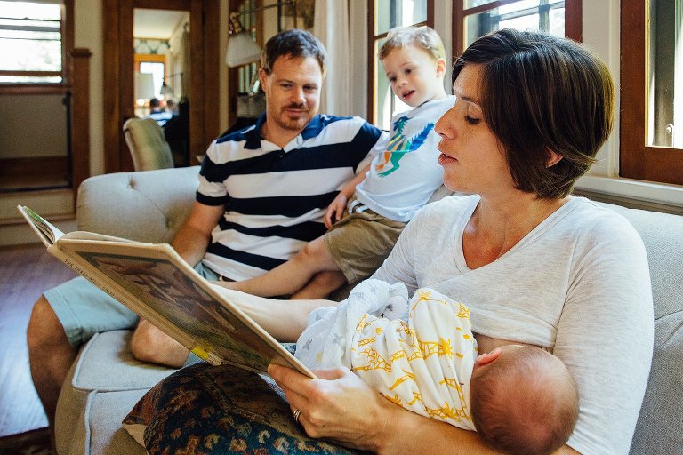 Mother breastfeeds newborn baby while reading a book to her son and husband.