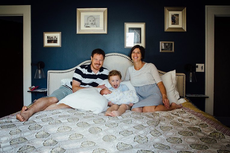 Family of four sits on a bed. Mom, dad, and young boy holding his newborn baby brother.