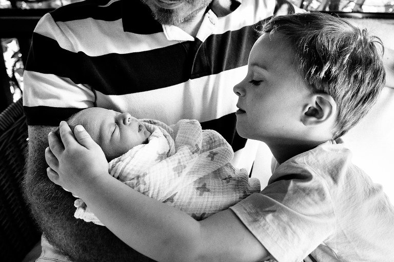Black and white. Father holds sleeping newborn baby. Young boy looks and touches his new sibling.