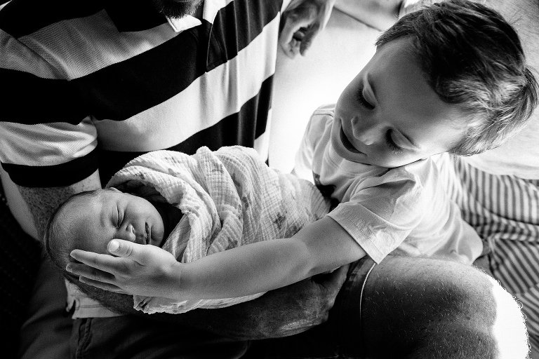 Black and white. Father holds newborn baby, older brother gently pats the baby's head.