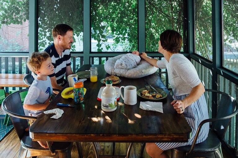 Family eating breakfast together all smile at their newborn baby sleeping on the table.
