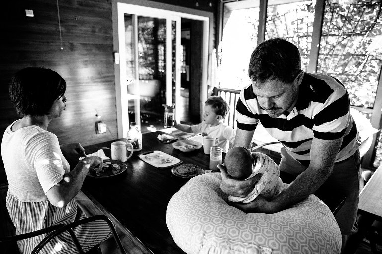 Black and white. Father picks up newborn baby as family eats breakfast on the back porch.