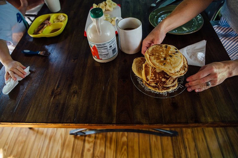 Family breakfast is served on the table. A large stack of chocolate chip pancakes, syrup, eggs, and a cup of coffee.