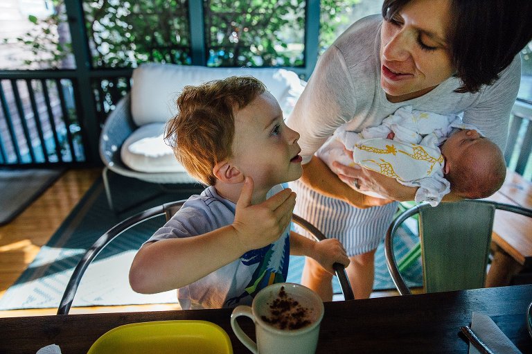 Mom holds newborn baby while son sits in the chair on the back porch with hot coca on the table.