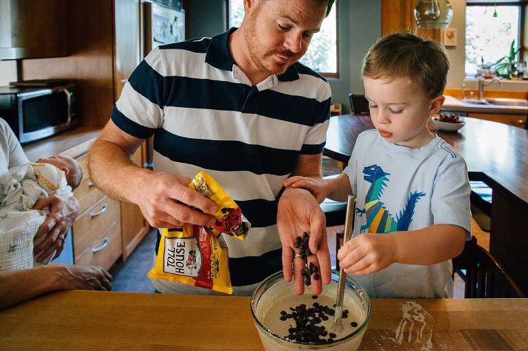 Dad helps his son pour chocolate chips into mixing bowl with pancake batter in it. Mother stands next to them holding sleeping newborn baby.