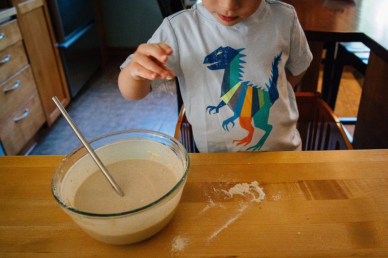 Pancake batter sitting in glass mixing bowl. Young boy standing on stool holds up his hand with some batter on it.
