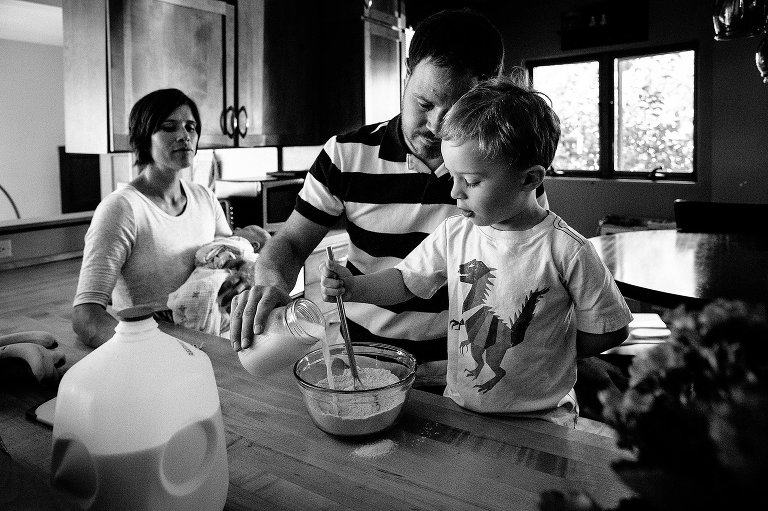 Black and white. Family making breakfast together in the kitchen. Mother stands holding newborn baby as father helps son pour in milk and stir to make pancake batter fro breakfast.