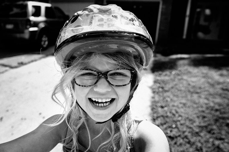 Young girl wearing her helmet standing outside on the driveway with a big smile on her face.