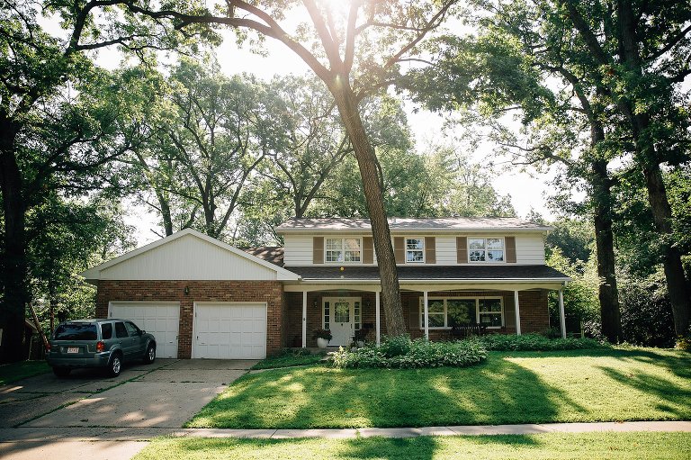 Traditional brick house with a car parked in the driveway and many large trees surrounding the house with the sun shining through.
