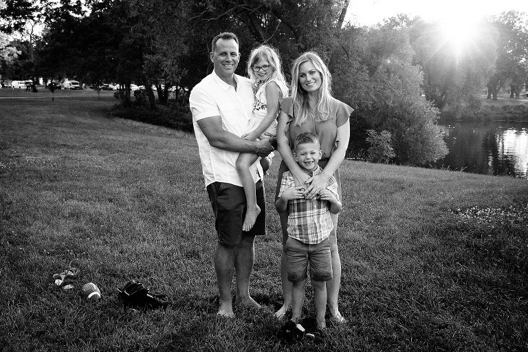 Black and white. Family of four standing in the grass next to the lake for a family picture. Sun setting behind them.