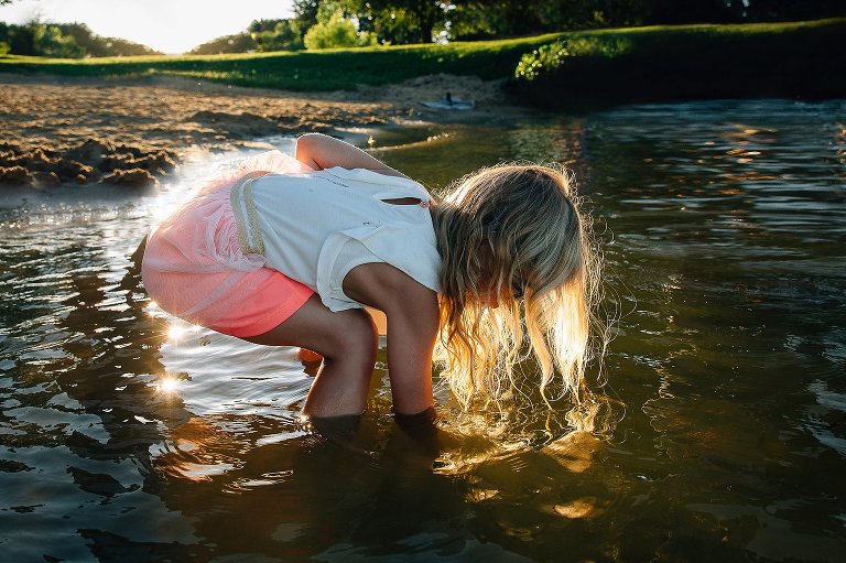Young girl standing in the water bends over with her hand in the water. The ends of her curly blonde hair fall into the water.