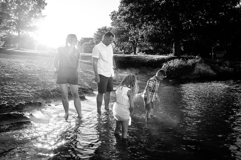 Black and white. Family of four standing at the beach with their feet in the water as the sun sets behind them.