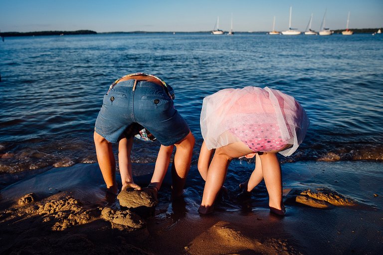 Brother and sister stand at the beach bent over both digging in the sand.